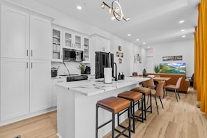 A modern kitchen with white cabinetry, a central island with stools, and a dining area with a table and chairs. - Hot Tub | 6BR-4BA | Sleep 12+ | Game Rm | Fire Pit (Louisville)