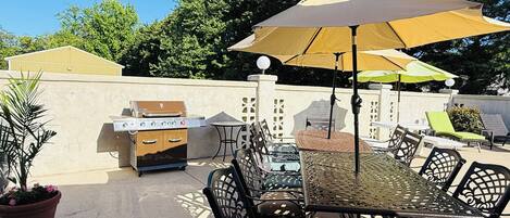 An outdoor patio with a grill, dining table, chairs, and umbrellas.
