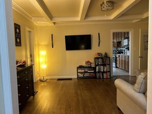 Una sala de estar con una televisión de pantalla plana, una estantería, un sofá y una cocina al fondo.