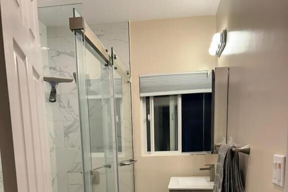 A bathroom with a glass shower enclosure, a sink, a mirror, and a window.