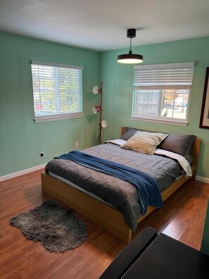 Un dormitorio con una cama, dos ventanas con persianas, una lámpara de noche y una alfombra pequeña.