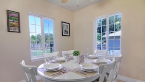A dining area with a round table set for four, white chairs, and a view of the backyard through large windows. - Spacious Room Prime Silicon valley (Palo Alto)