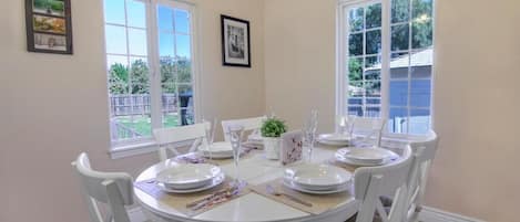 Un comedor con una mesa redonda dispuesta para cuatro personas, sillas blancas y vistas al jardín trasero a través de amplios ventanales.