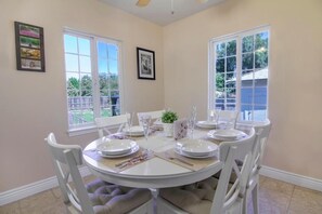 A dining area with a round table set for four, white chairs, and a view of the backyard through large windows. - Spacious Room Prime Silicon valley (PALO ALTO)