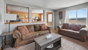 A living room with a brown leather sofa, a wooden coffee table, and a view of a kitchen and landscape through a large window. - Trapper 1 (West Yellowstone)