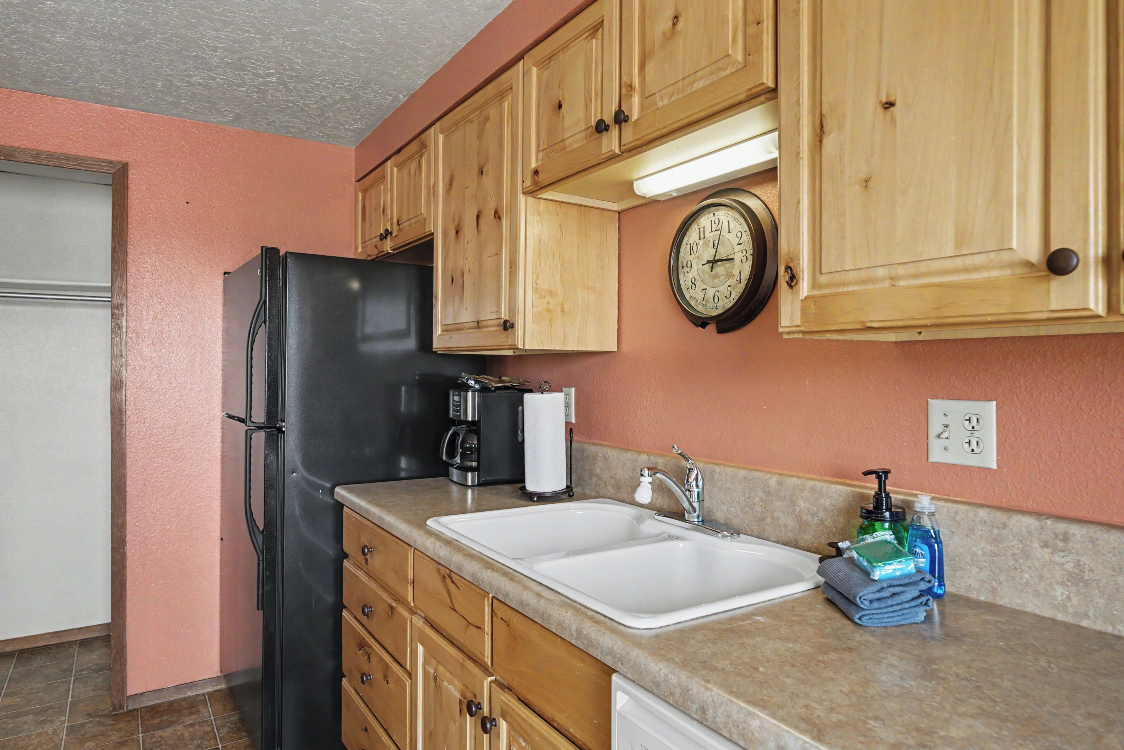 A kitchen with wooden cabinets, a black refrigerator, a coffee maker, a sink, and a clock on the wall.