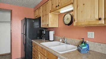 A kitchen with wooden cabinets, a black refrigerator, a coffee maker, a sink, and a clock on the wall.