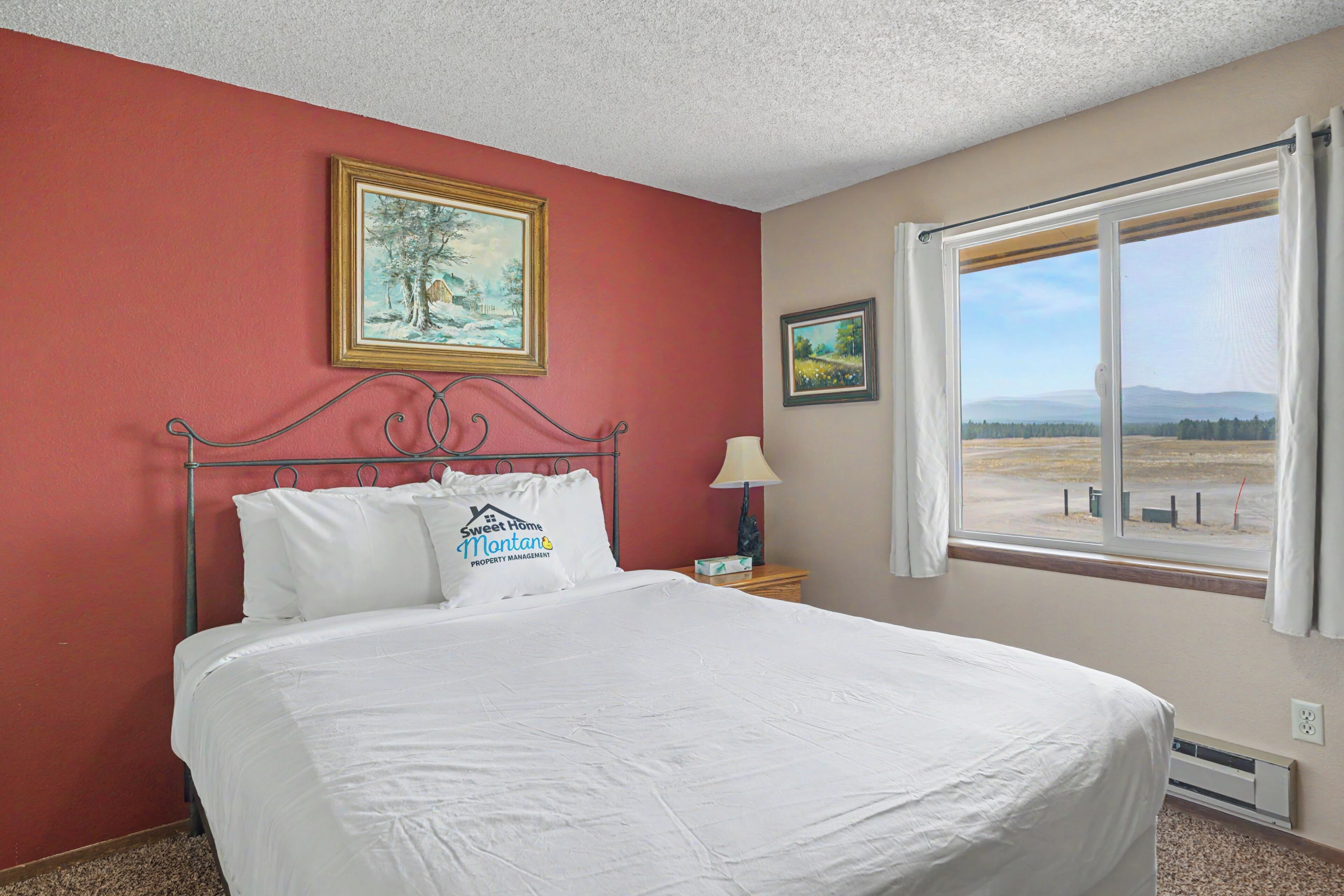 Habitación de hotel con una cama grande, dos cuadros en la pared, un ventanal con vistas y una señal en la almohada.