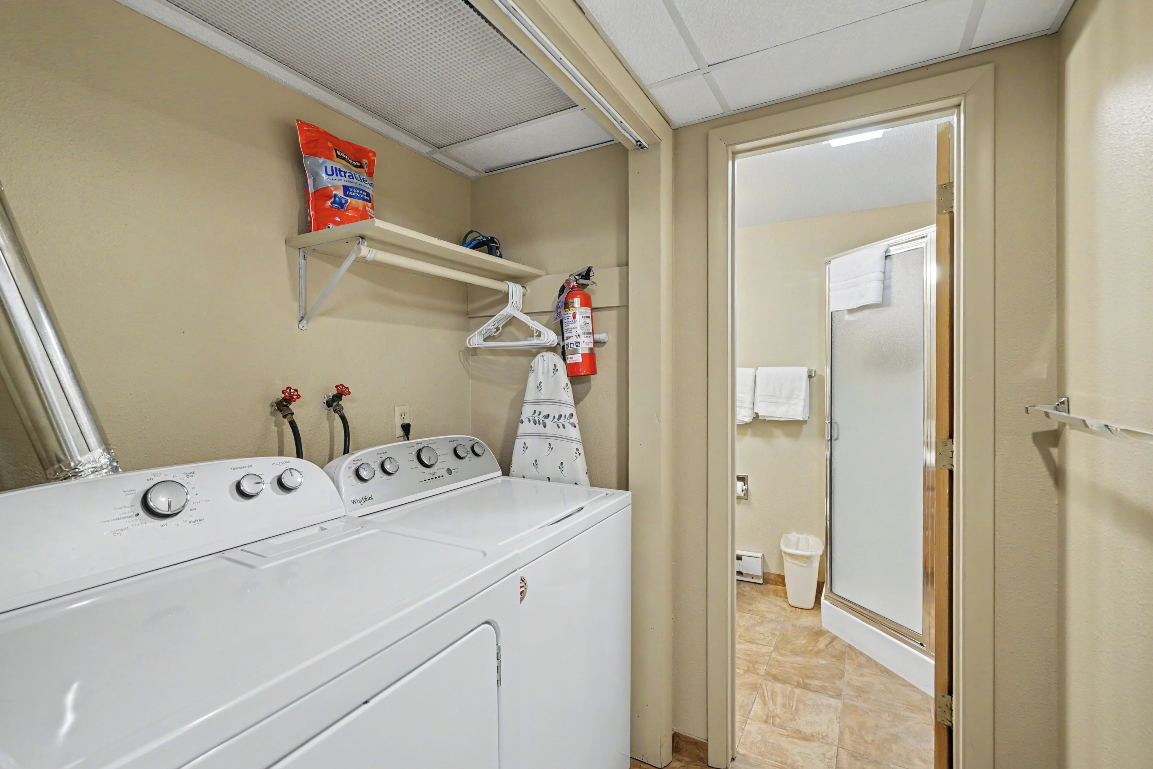 A laundry room with a front-loading washer and dryer, a fire extinguisher, and a shelf above the dryer.