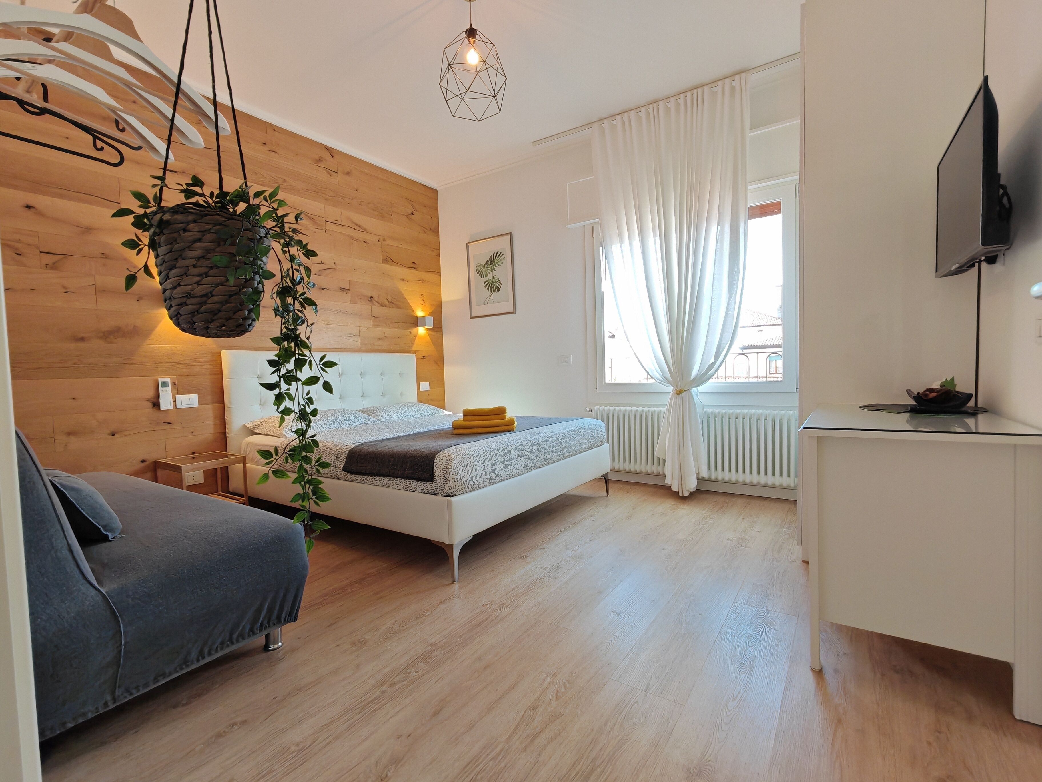 Une chambre moderne avec un mur en bois, un lit doté d’une tête de lit blanche, une table de chevet, une plante suspendue et une fenêtre avec des rideaux.