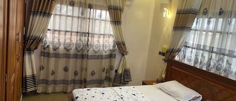 A hotel room with a bed, curtains, an air conditioning unit, and a nightstand.