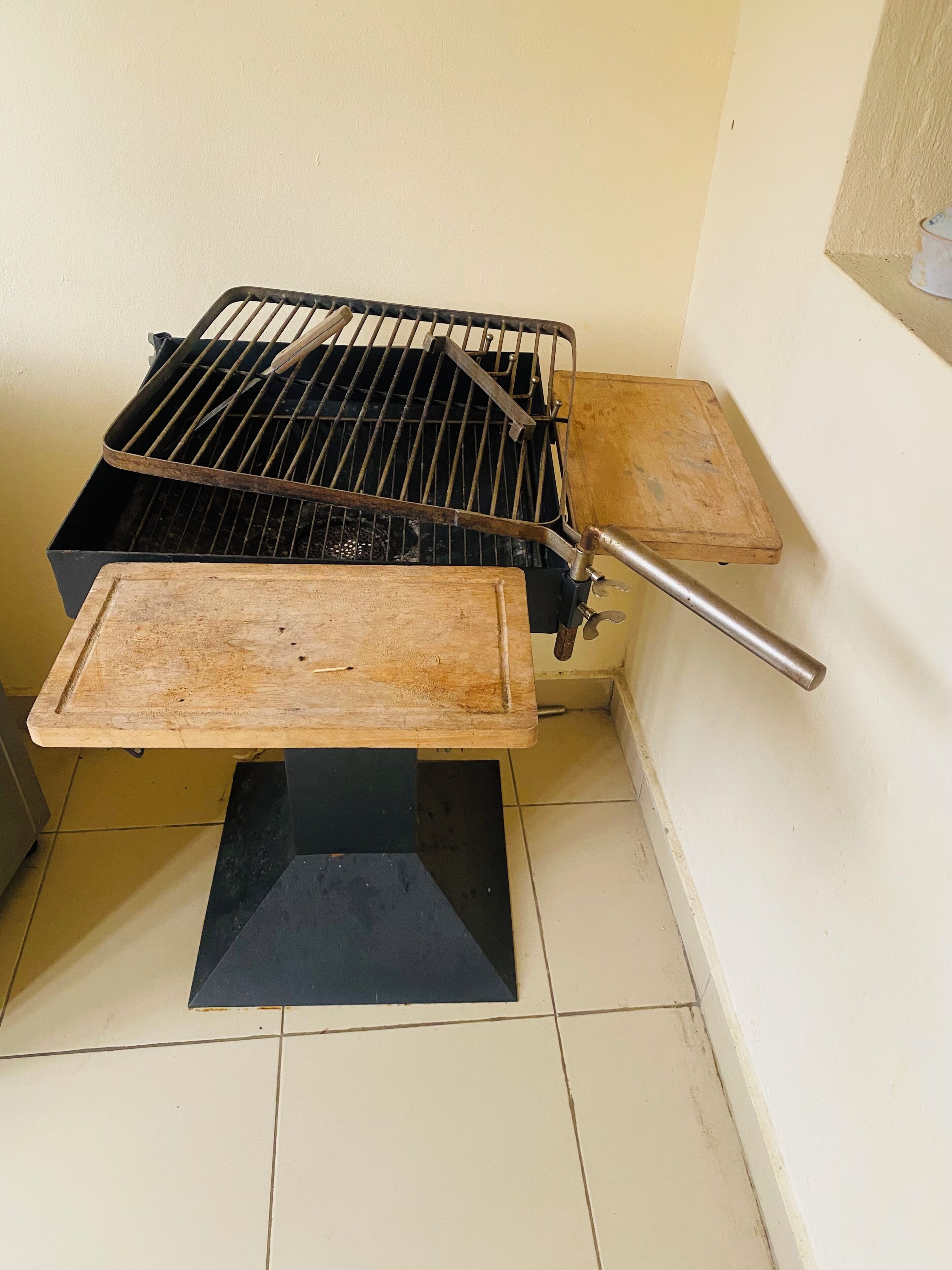 Ein tragbarer Grill mit einem Metallrost und einer Holzablage, auf einem Fliesenboden vor einer Wand platziert.