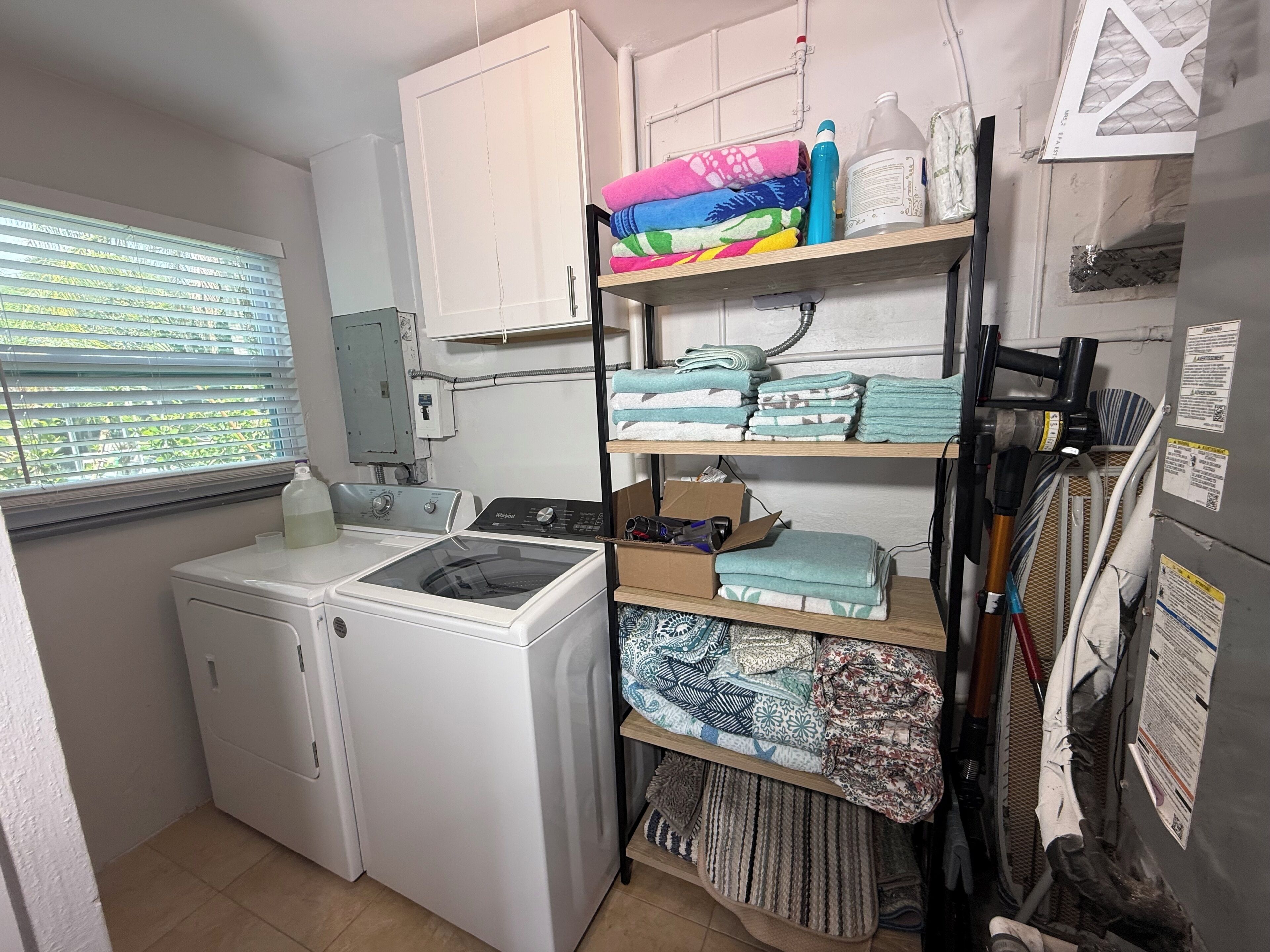 A laundry room with a front-loading washer and dryer, shelves with folded towels, and a window with blinds.