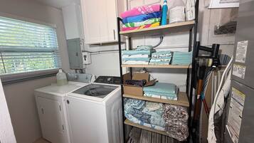 A laundry room with a front-loading washer and dryer, shelves with folded towels, and a window with blinds.