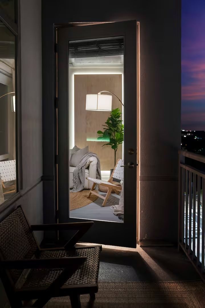 Un balcón con una silla y una mesita, una puerta que da a un cuarto con una lámpara y una planta, y vista al cielo nocturno.