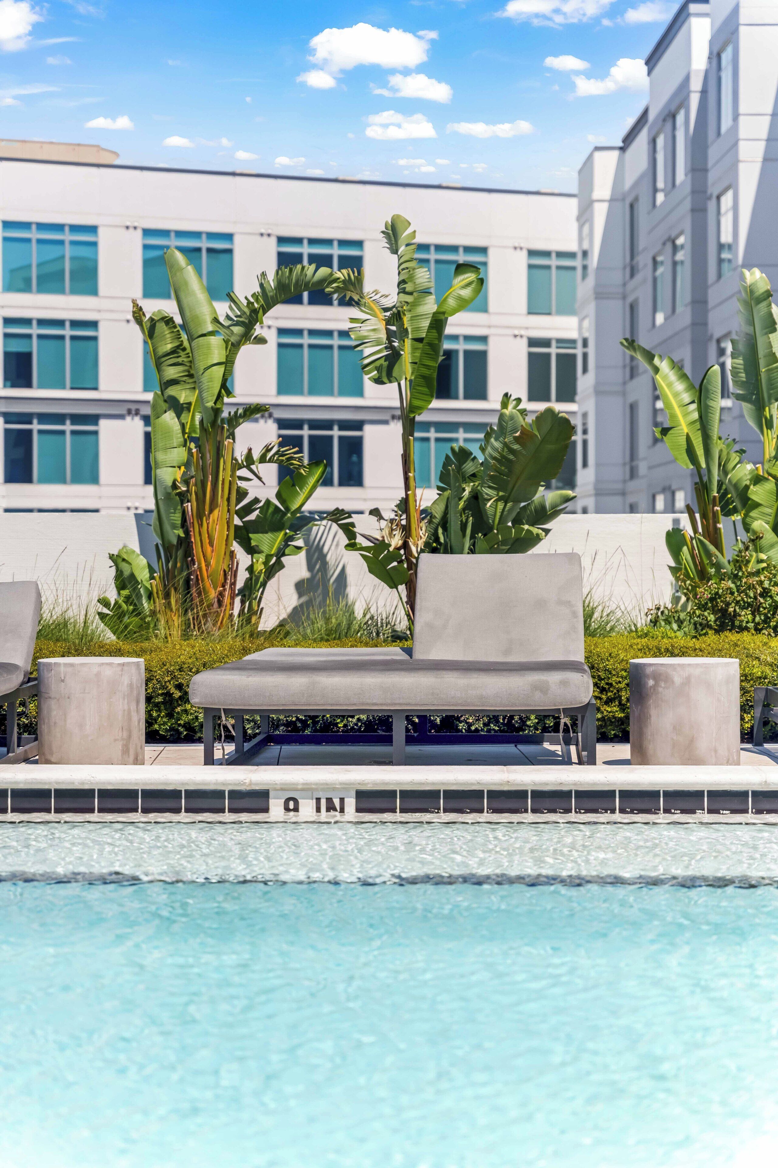 Ein Liegebereich am Pool mit Liegestuhl und kleinem Tisch, umgeben von tropischen Pflanzen und einem modernen Gebäude im Hintergrund.
