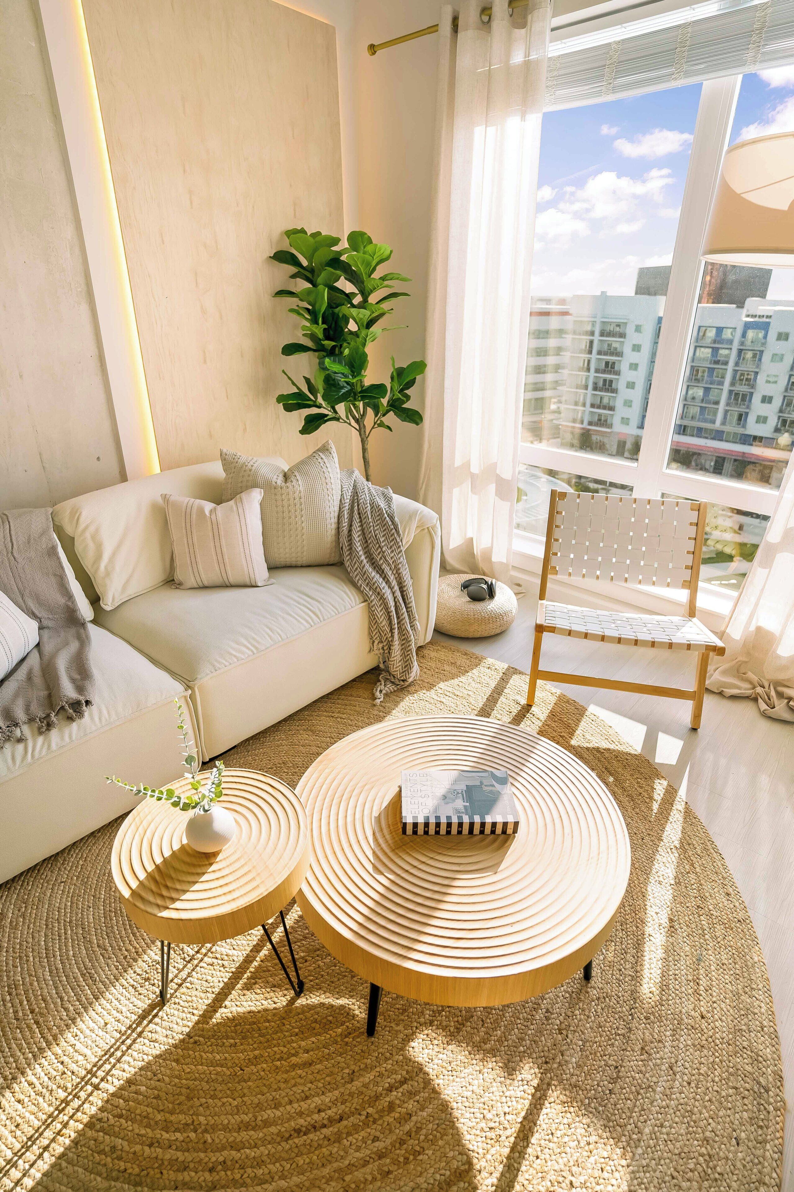 A modern living room with a sofa, two round wooden tables, a chair, and a large window with a view of buildings.