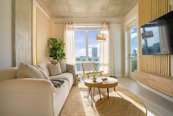 A modern living room with a sofa, a coffee table, a plant, and a view of the city through large windows. - The Emma - Luxury 1BR Condo King Bed, Pool, Gym (Orlando)