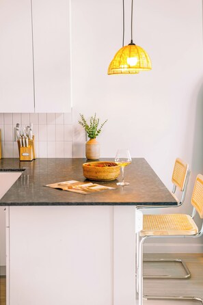 A kitchen with a central island, a hanging pendant light, a vase with greenery, a wine glass, and a basket of fruit. - The Emma - Luxury 1BR Condo King Bed, Pool, Gym (Orlando)