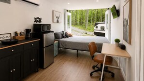 Et lille værelse med tekøkken, seng, skrivebord og et vindue med udsigt til skov og en autocamper.