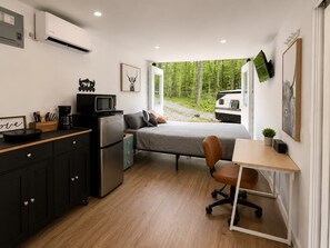 Ein gemütlicher Raum mit Küchenzeile, einem Bett, einem Schreibtisch und einem Fenster mit Blick auf einen Wald und einen Wohnwagen.