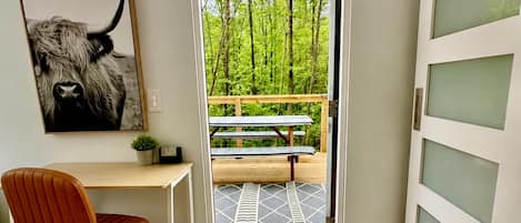 A room with a desk, chair, and a large window leading to a balcony with a bench.