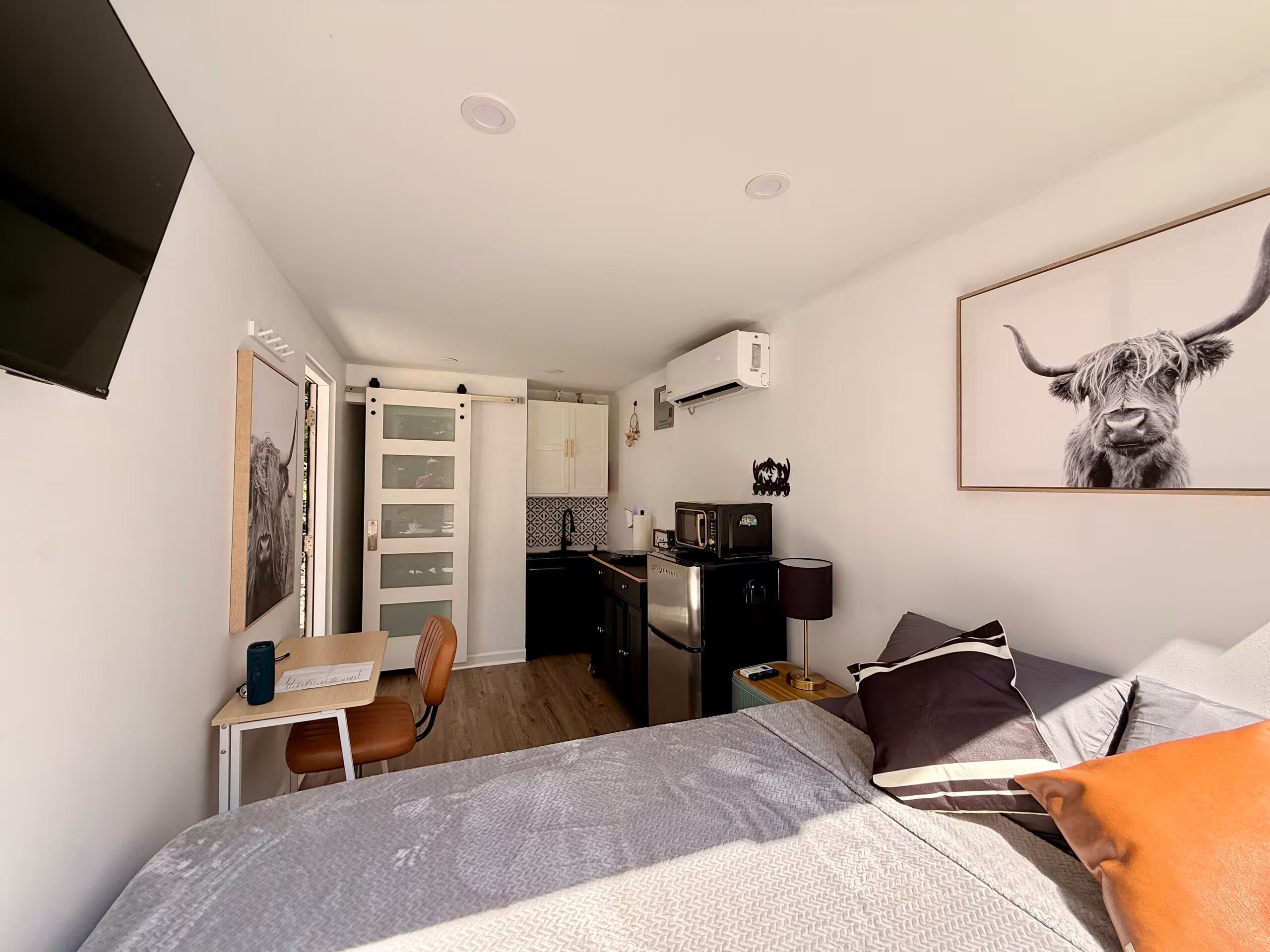 Habitación compacta con cama, escritorio, silla, kitchenette y una obra de arte colgada.