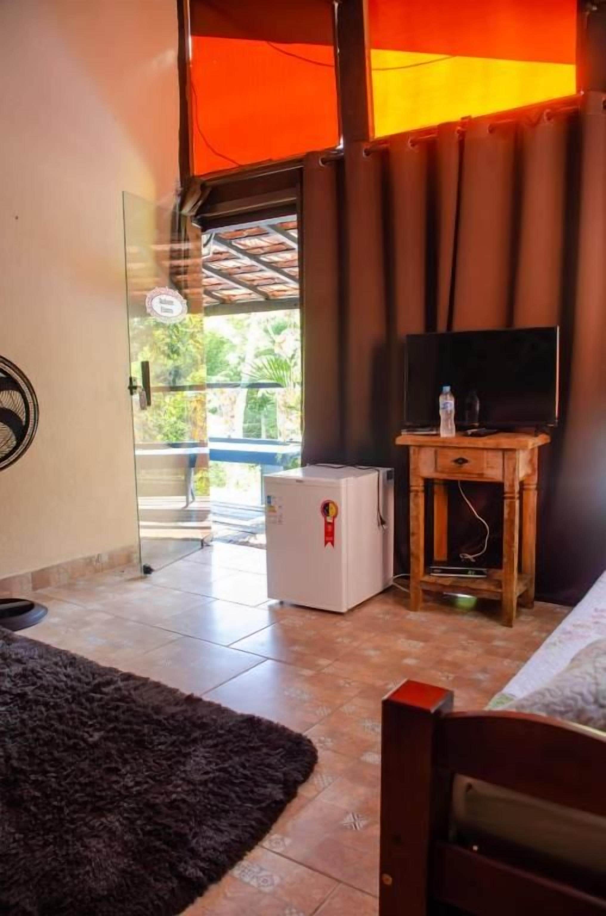 Una habitación con una cama, un televisor sobre un mueble de madera, un refrigerador pequeño y una puerta de vidrio que da al exterior.