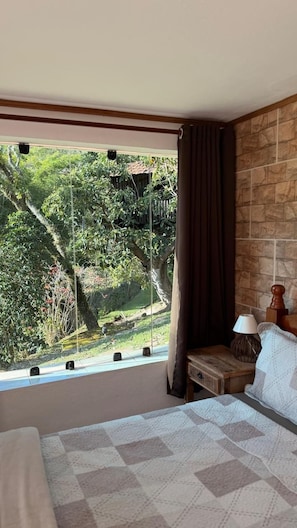 A bedroom with a large window, a bed with a patterned bedspread, a nightstand with a lamp, and a view of greenery outside. - Pousada Estância Vip (Cunha)