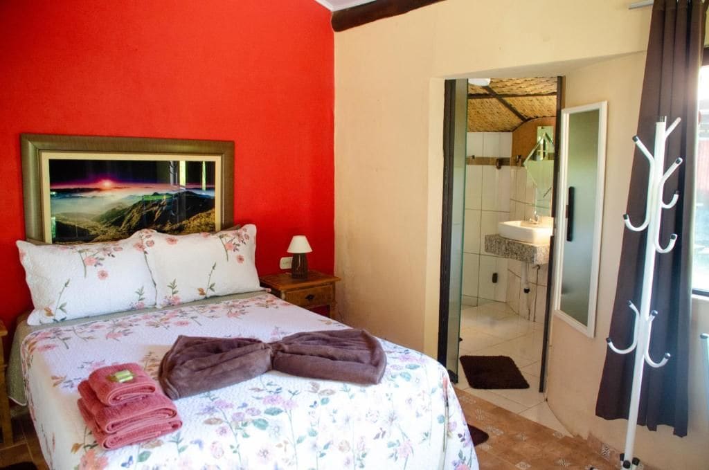 A bedroom with a bed, a painting, a bathroom visible through an open door, and a coat rack.