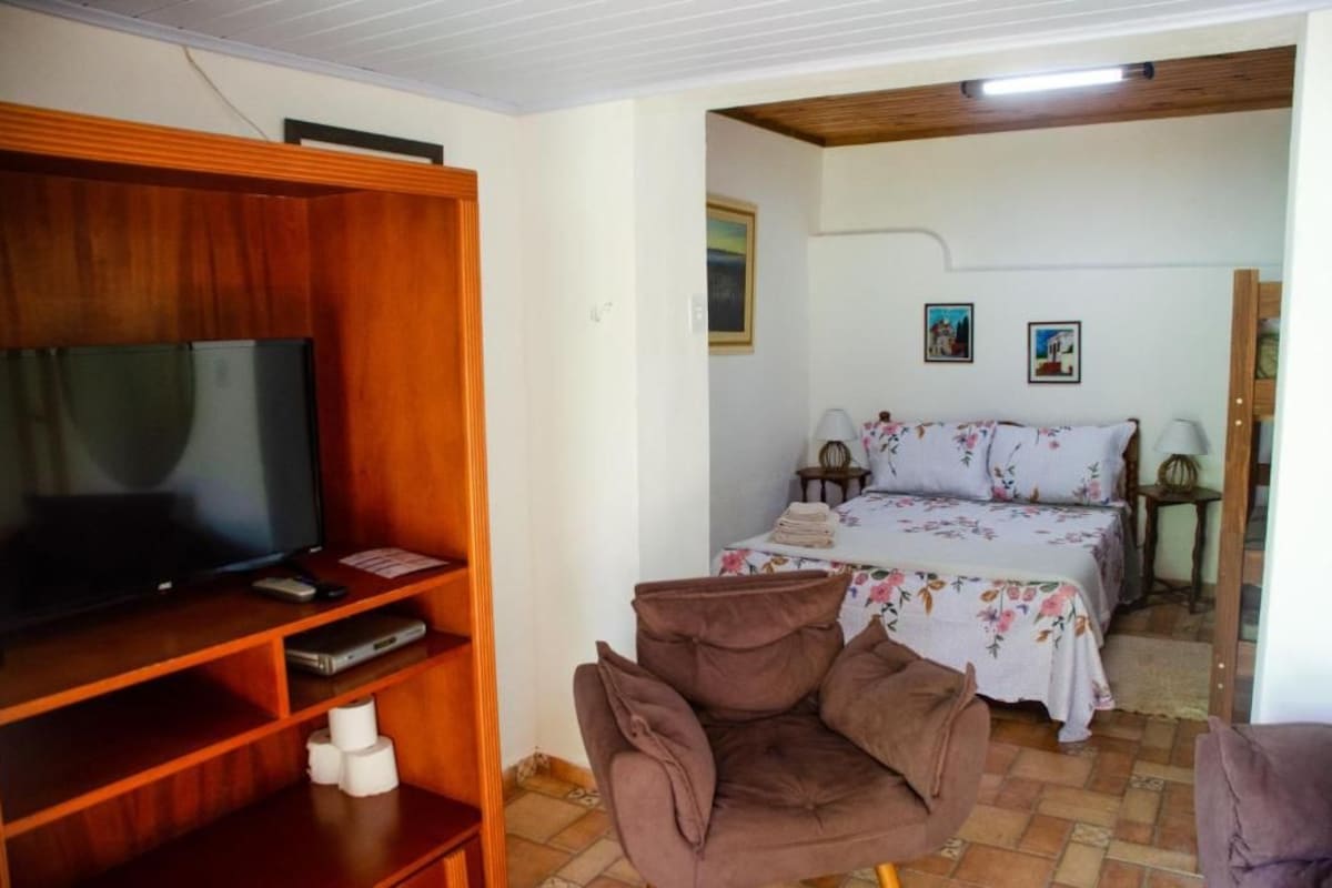 Habitació amb televisió sobre un suport de fusta, un llit amb llençols florals i una butaca marró.