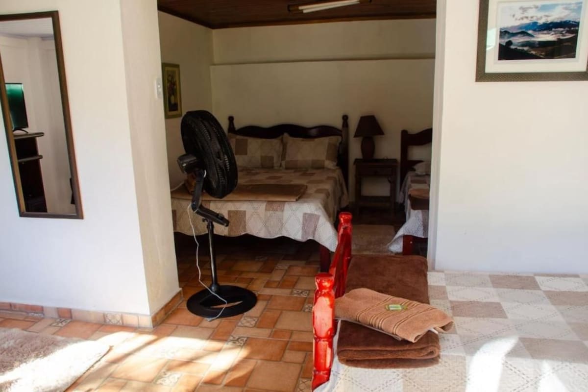 Un dormitori amb un llit, un ventilador de peu, una cadira i una taula petita.