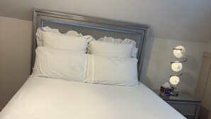 Una cama bien tendida con cabecera, una mesita de noche con lámpara, y una lámpara de pared.