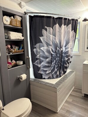 A bathroom with a toilet, a shower curtain with a floral design, a shelf with toiletries, and a towel rack. - 2-bedroom bed & breakfast in nice Syracuse with front fenced in yard (Syracuse)