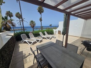 Eine Terrasse mit Tisch und Stühlen, einem Grill und Liegestühlen mit Blick auf das Meer und Palmen.
