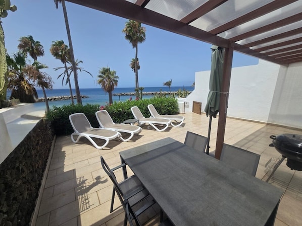A patio with a table and chairs, a grill, and lounge chairs facing a view of the sea and palm trees. - 2 Bedroom Beachfront Playa Flamingo Apartment with Private Terrace & Air Con (Playa Blanca)