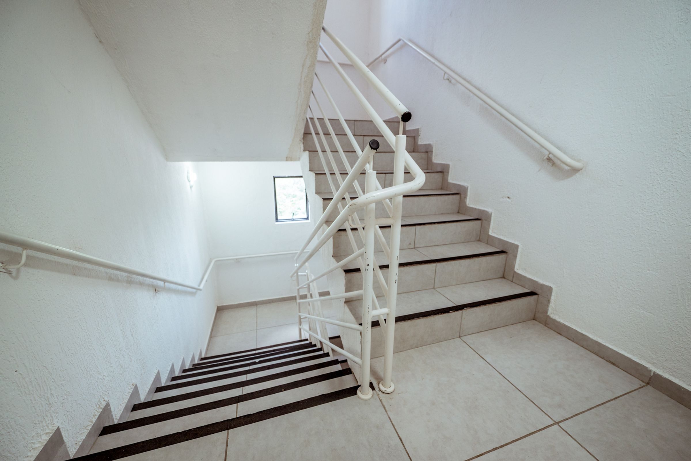 Uma escadaria com corrimão, degraus azulejados e uma pequena janela no topo.