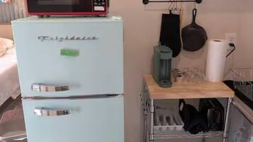 A compact kitchen with a light blue Frigidaire refrigerator, a microwave on top, a small cart with kitchen items, and a hanging pot rack.
