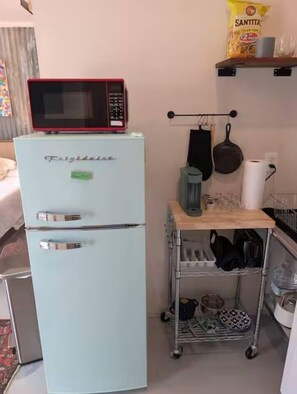 A compact kitchen with a light blue Frigidaire refrigerator, a microwave on top, a small cart with kitchen items, and a hanging pot rack.