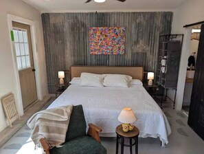 A bedroom with a large bed, two bedside lamps, a colorful abstract painting, a radiator, and a chair.