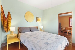 A bedroom with a bed, two bedside tables, a wall-mounted round mirror, and a door leading to a living area.