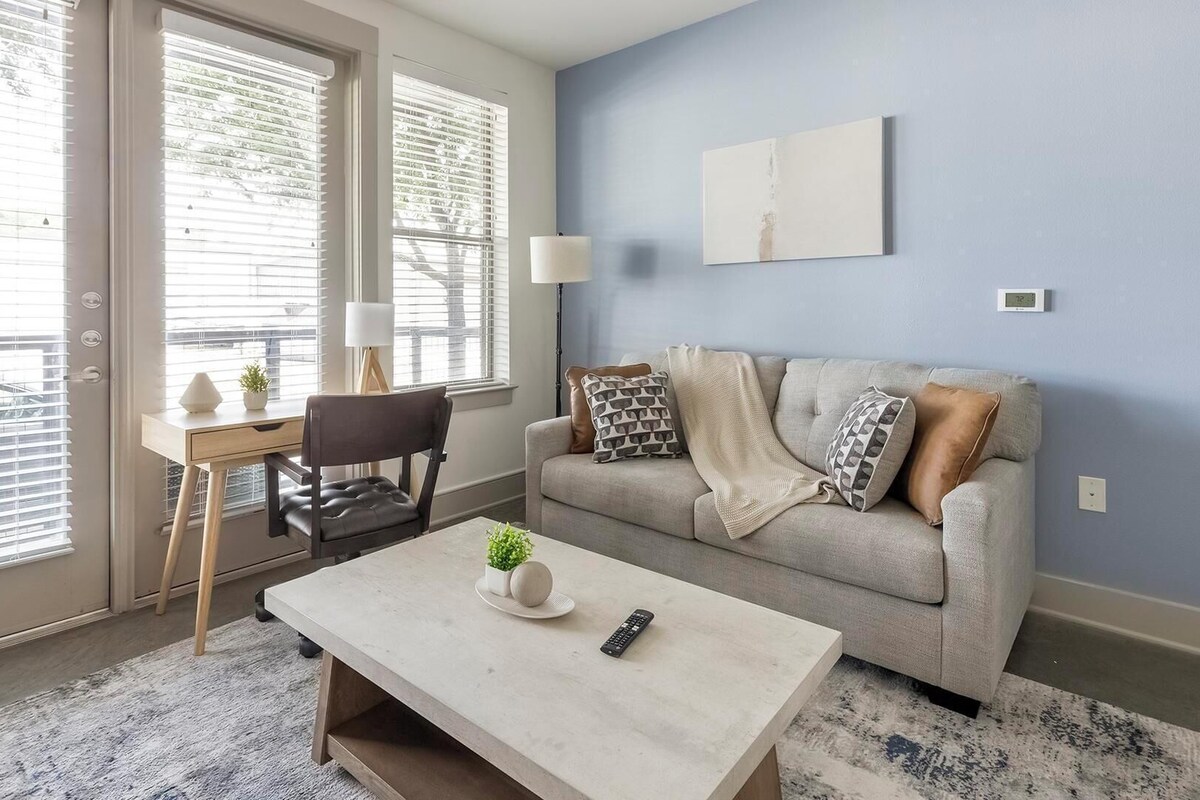 Ein Wohnzimmer mit einer Couch, einem Couchtisch und einem Sessel. Ein Fenster mit Jalousien, eine Lampe und eine Wanduhr sind ebenfalls vorhanden.