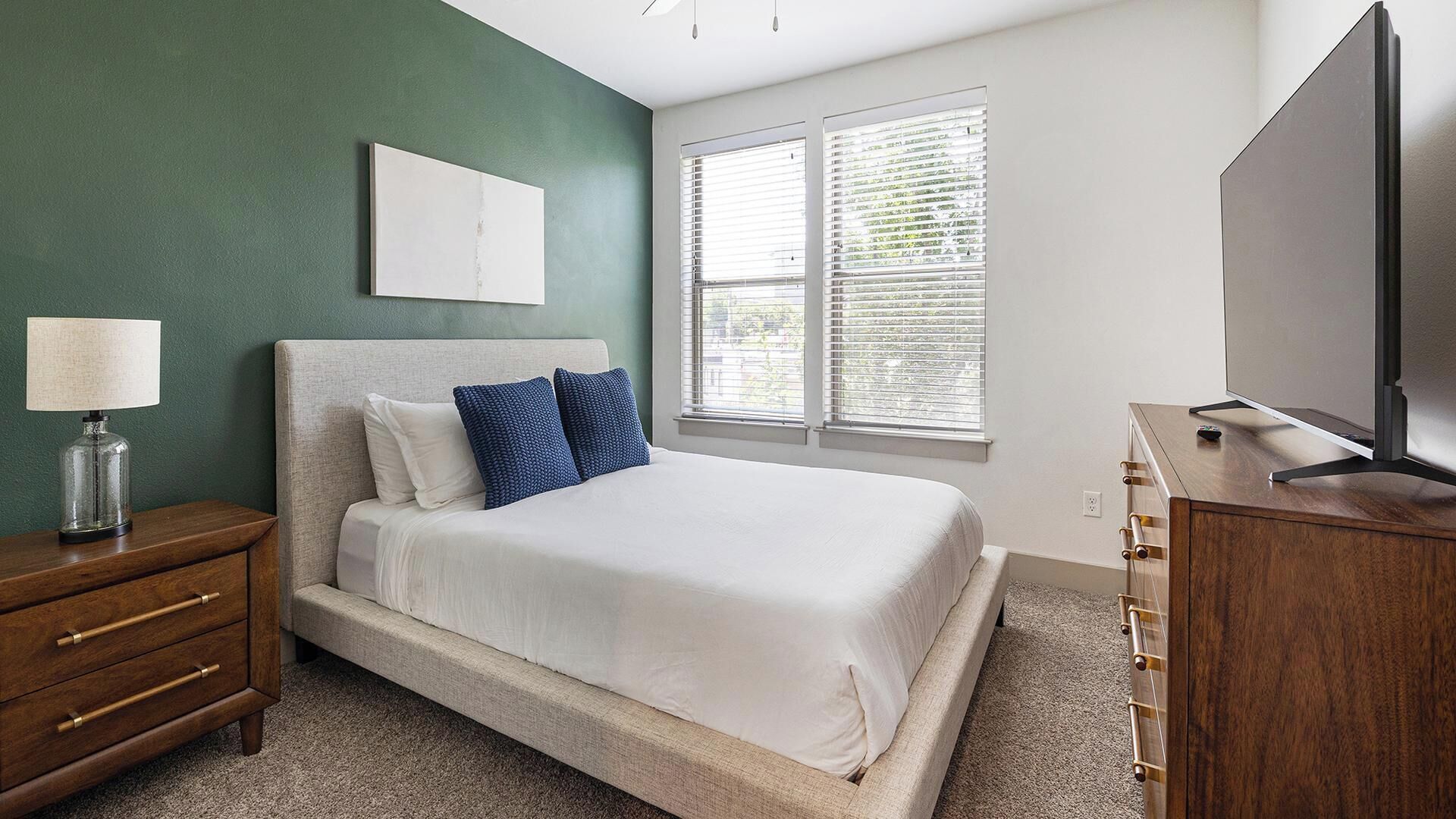 A bedroom with a bed, a nightstand, a dresser, a TV, and a window with blinds.