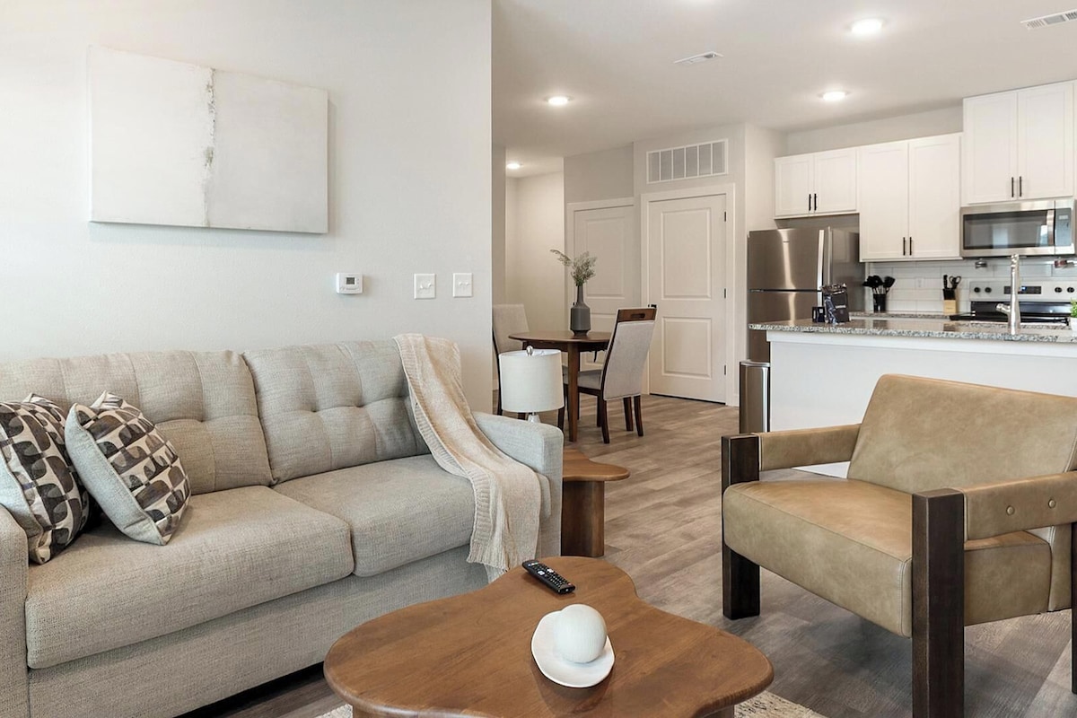 A living room with a sofa, armchair, and coffee table. There is a kitchen in the background with modern appliances.