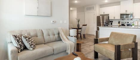 Una sala de estar con un sofá, un sillón y una mesa de centro. Se observa una cocina al fondo con electrodomésticos modernos.