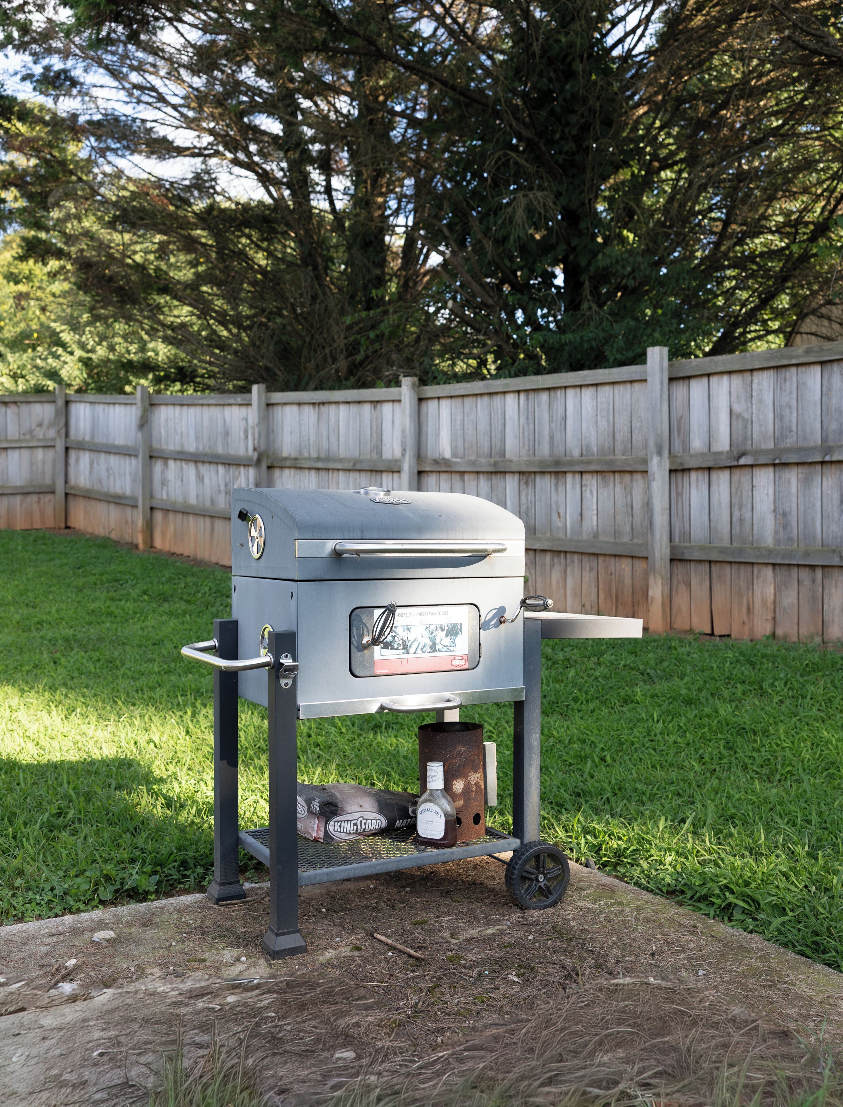 A portable barbecue grill with a metal grate, a handle, and a lid, placed on a grassy area with a wooden fence in the background.