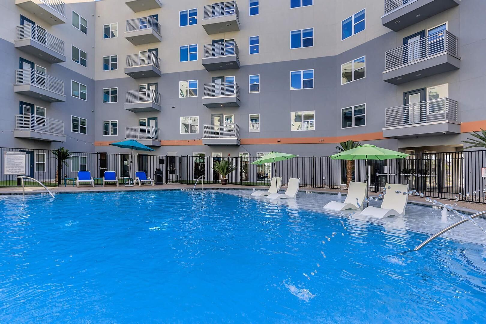 Ein Swimmingpool mit Liegestühlen und Sonnenschirmen, umgeben von einem mehrstöckigen Gebäude mit Balkonen.