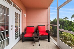 A balcony with two wicker chairs and a small table, a white door with glass panels, and a view of greenery. - Seaside At Anastrasia A102 Go Coastal (St. Augustine)