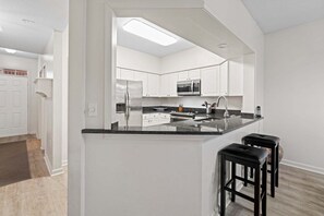 A modern kitchen with white cabinets, a stainless steel refrigerator, a microwave, and a black countertop. There are three black bar stools at the counter. - Seaside At Anastrasia A102 Go Coastal (St. Augustine)