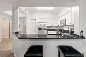 A modern kitchen with a central island, white cabinetry, stainless steel appliances, and a black countertop. - Seaside At Anastrasia A102 Go Coastal (St. Augustine)